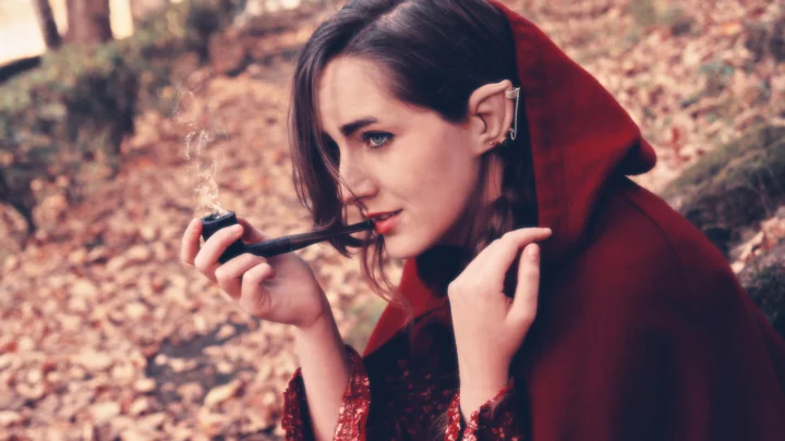 一名成年女子在落叶覆盖的户外环境中披着红色兜帽斗篷并叼着烟斗。