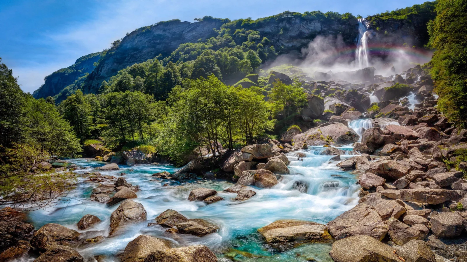 A sunlit waterfall cascades through a forested mountain landscape into a clear turquoise stream flowing over rocky terrain.