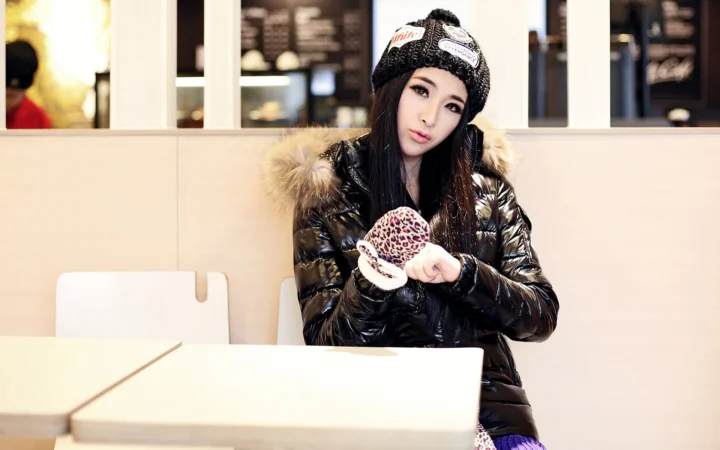 一名戴针织帽、穿黑色冬季外套的女性坐在室内桌旁。