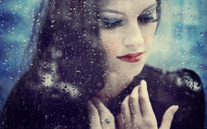一名女性隔着布满雨滴的玻璃闭眼微笑，画面呈现蓝绿色调的近距离肖像。