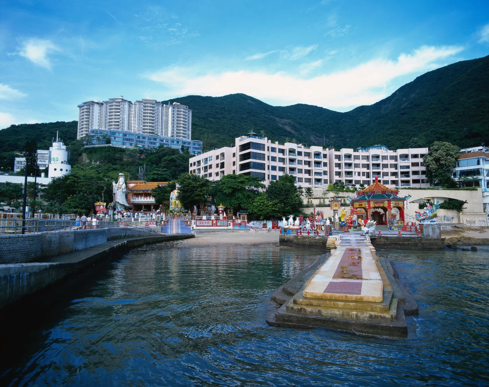 一座滨水寺庙坐落在高楼与青山之间，前方水面倒映着建筑和天空。