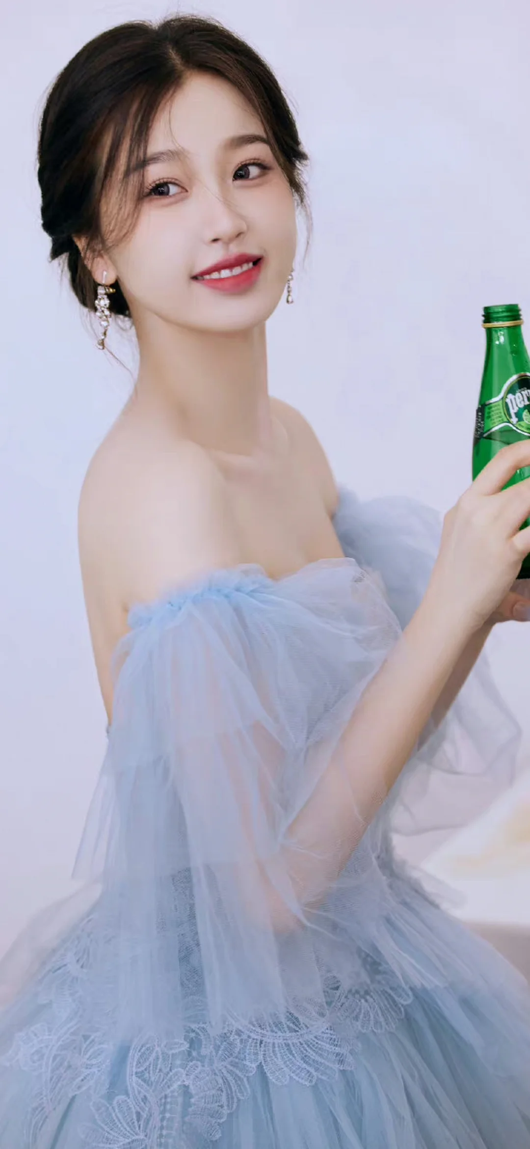 一名成年女子身穿淺藍色禮服，手持綠色瓶子面對鏡頭。