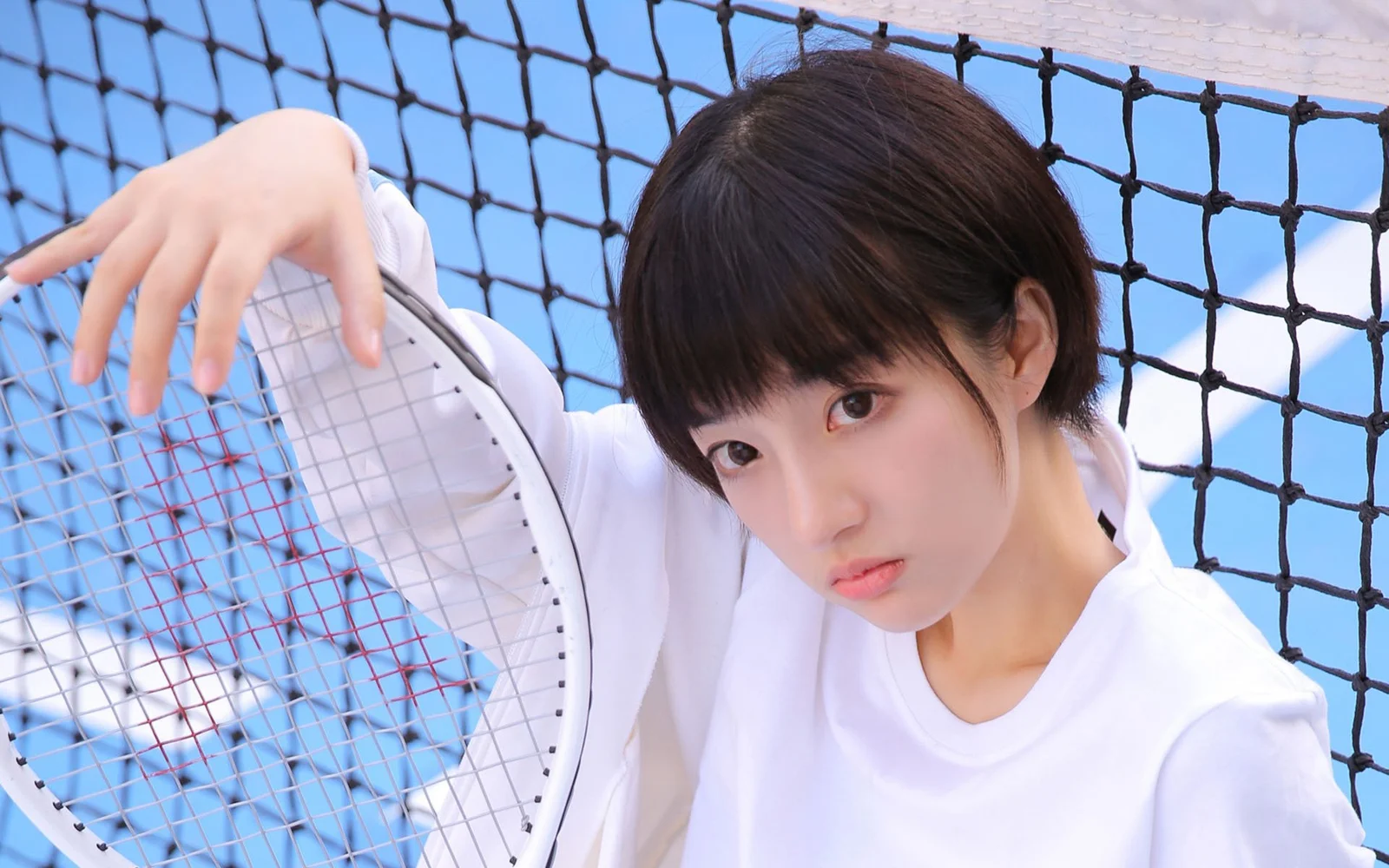 白いトップスの女性がテニスラケットを持ってテニスネットの前に立っています。