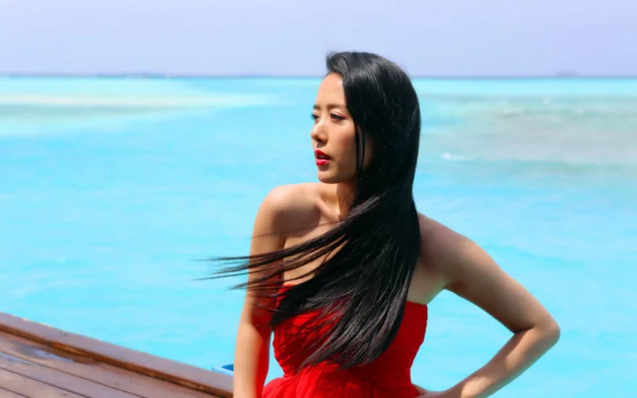 一名身穿红裙的成年女子站在明亮的蓝色海水前。