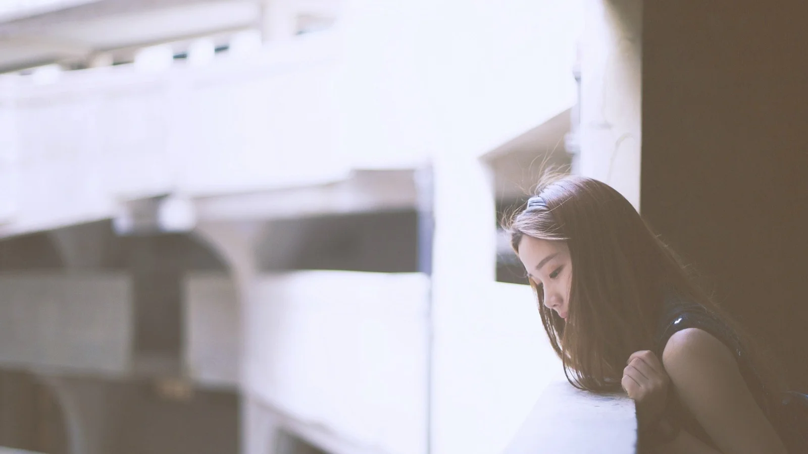 긴 머리의 여성이 밝은 흰색 건축 구조물 옆에 기대어 있는 부드러운 색조의 이미지입니다.