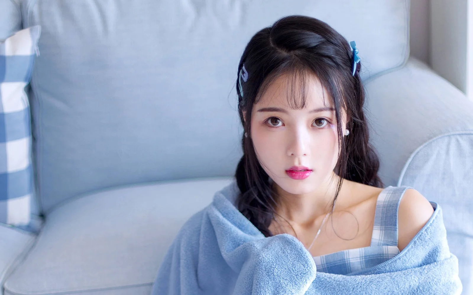 一名深色长发女性裹着浅蓝色毯子坐在蓝色沙发上面对镜头。