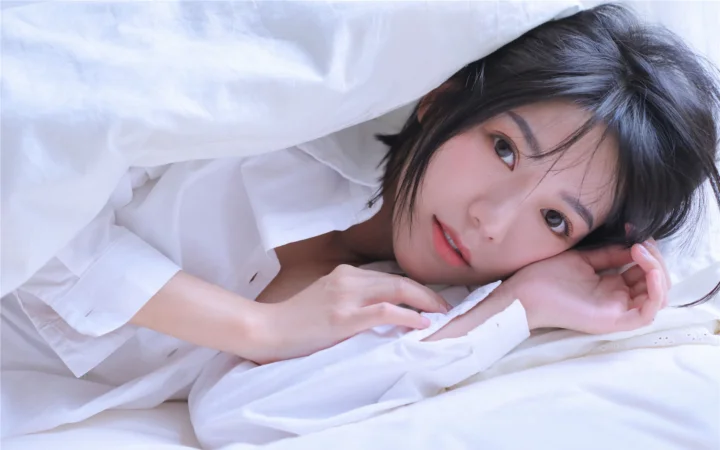 一名身穿白色衬衫的成年女子侧躺在白色床品上看向镜头。