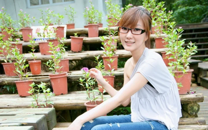一名戴眼镜的成年女性坐在摆满盆栽的户外木架旁。