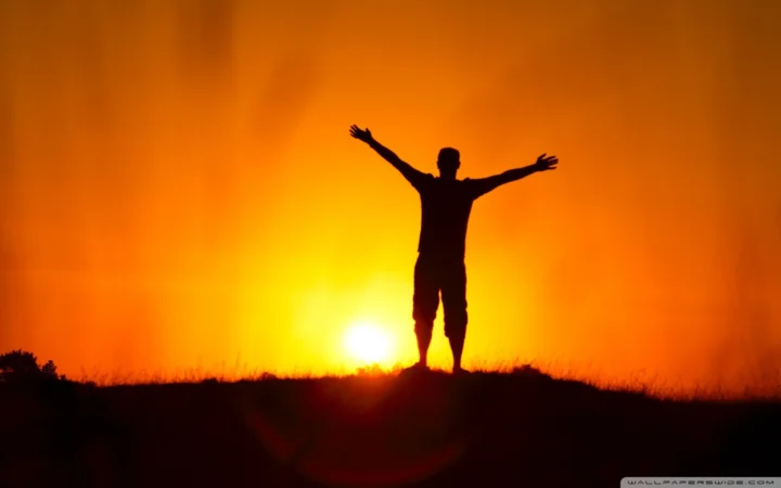 橙色夕阳映照下，一名张开双臂的人站在地平线上形成剪影。