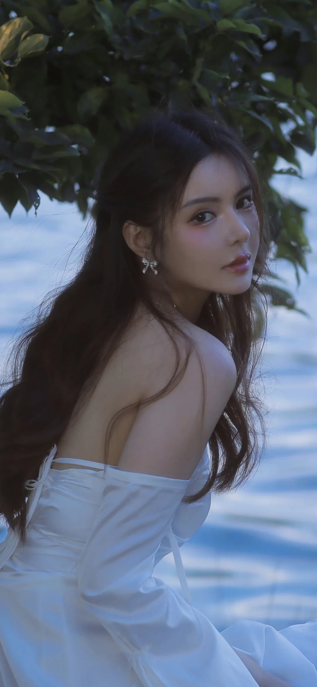 一名长发女性穿着白色露肩服装坐在水边，在树叶掩映下回头望向镜头。