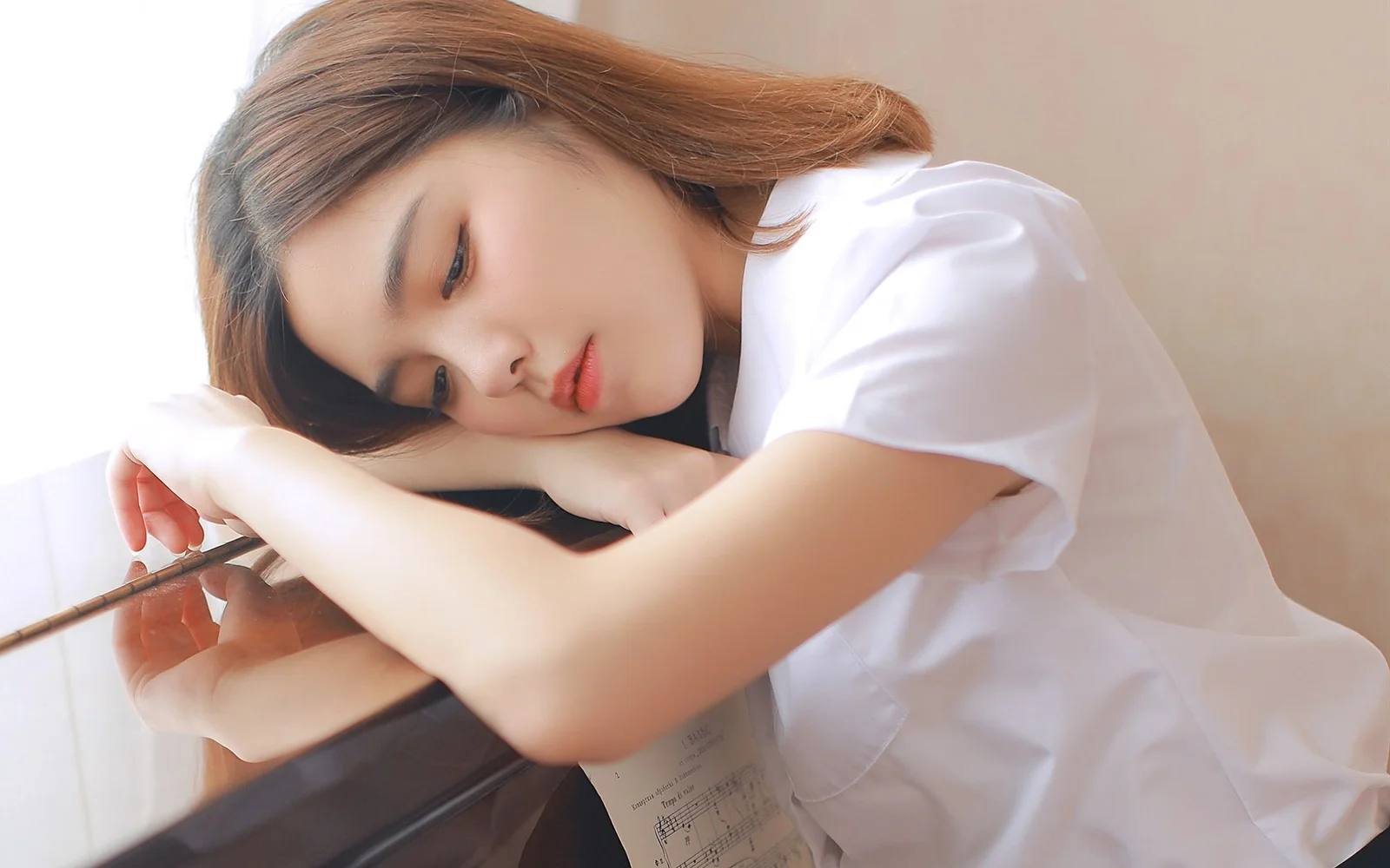 흰색 상의를 입은 여성이 피아노에 옆으로 기대어 차분하고 부드러운 표정을 짓고 있다.