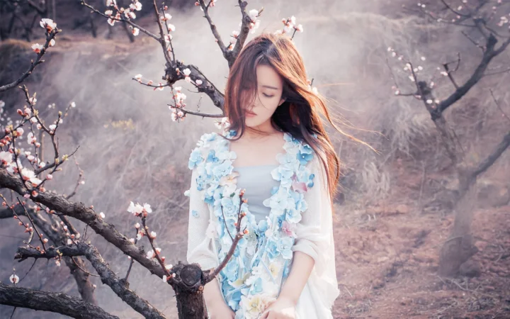 一名身穿浅蓝色连衣裙的女子站在盛开梅花的树枝间。