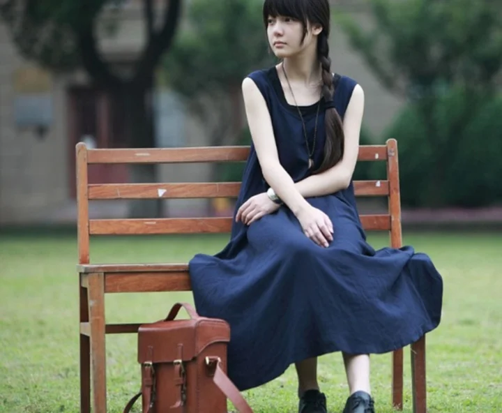 濃紺のワンピースを着た女性が、緑の公園にある木製ベンチに座り、隣に茶色の革バッグが置かれています。