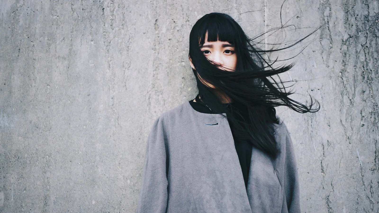 灰色のコートを着た黒髪の女性が、風に長い髪をなびかせながら灰色の質感のある壁の前に立っています。