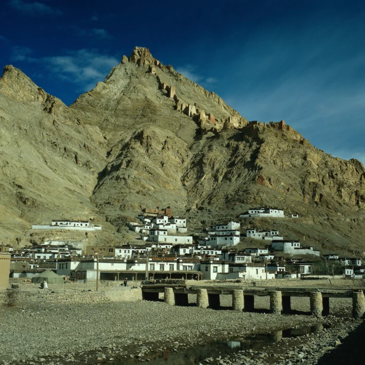 陡峭的岩山下坐落着一片白色建筑村落，前方可见横跨溪流的石桥。