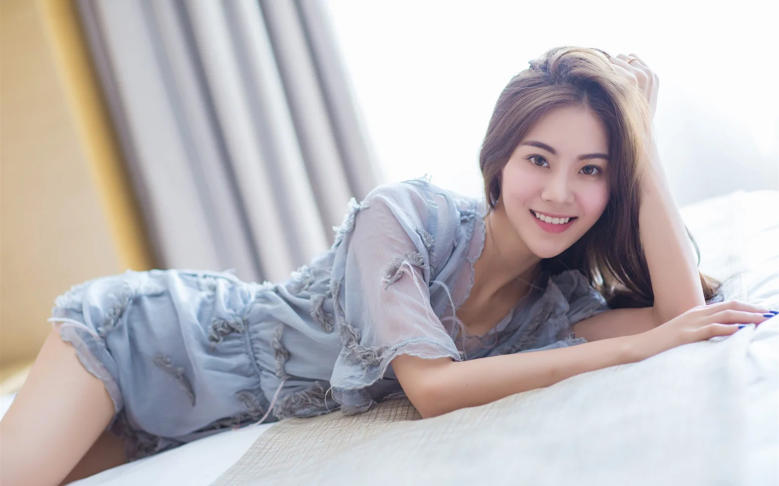 一名成年女性穿着浅蓝色连衣裙，微笑着侧躺在窗边的床上。