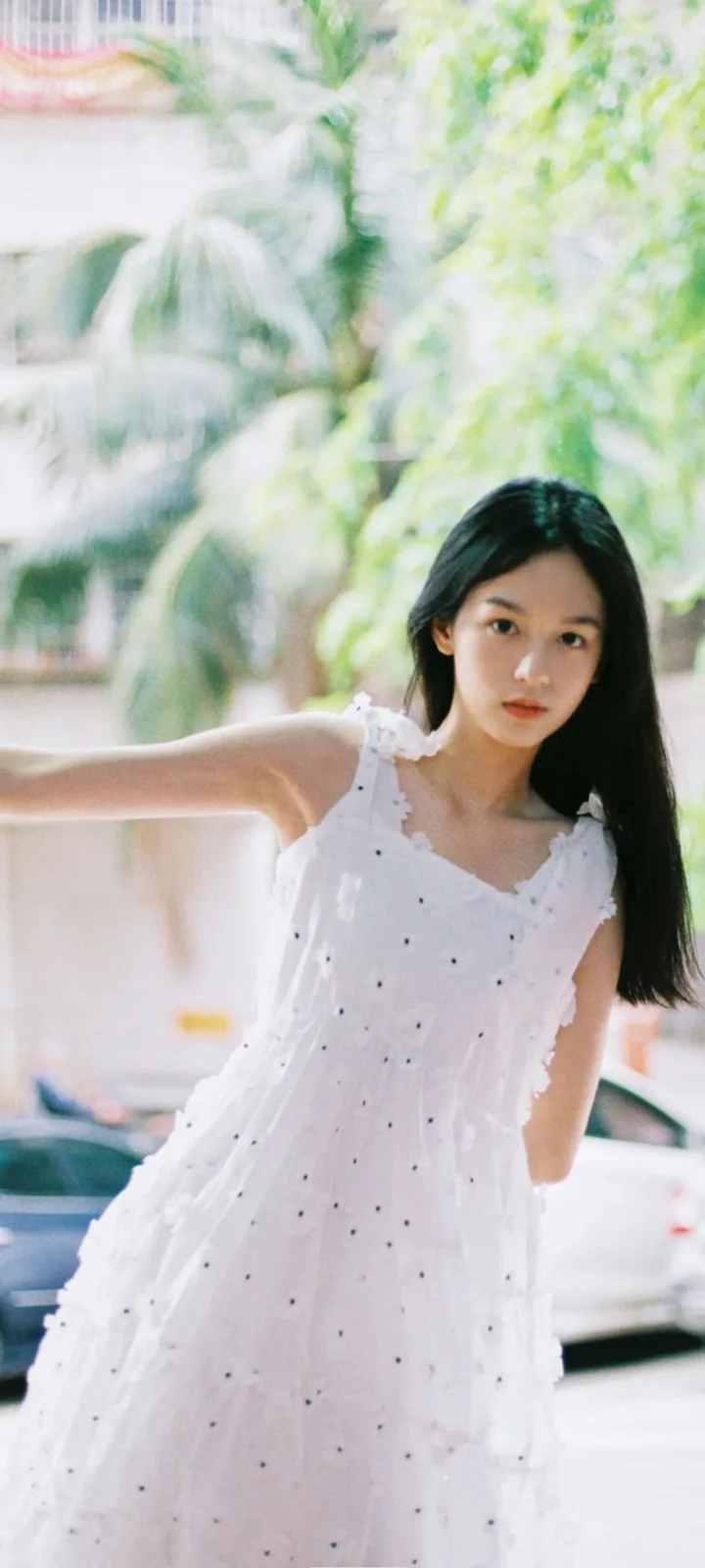 一名长发女子穿着白色连衣裙站在绿树和街道背景前。