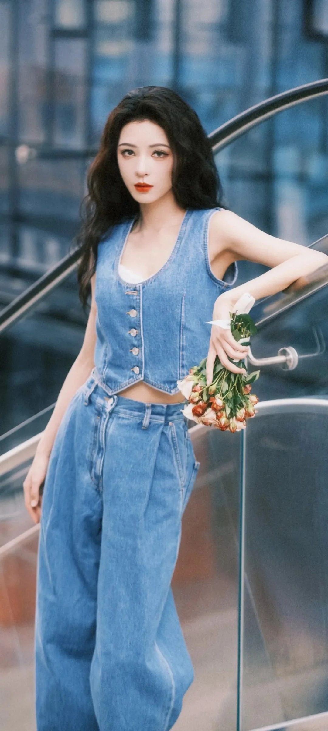 一名长发女子穿着蓝色牛仔背心和牛仔裤，站在扶梯旁手持一束小花。