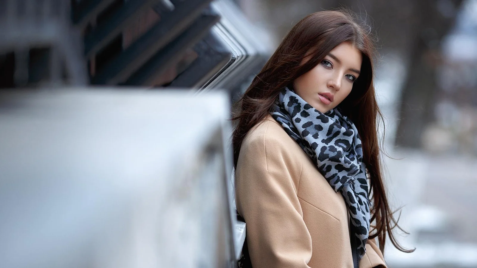 成人女性が車のそばに立ち、明るい色のコートとレオパード柄のスカーフを身に着けている。