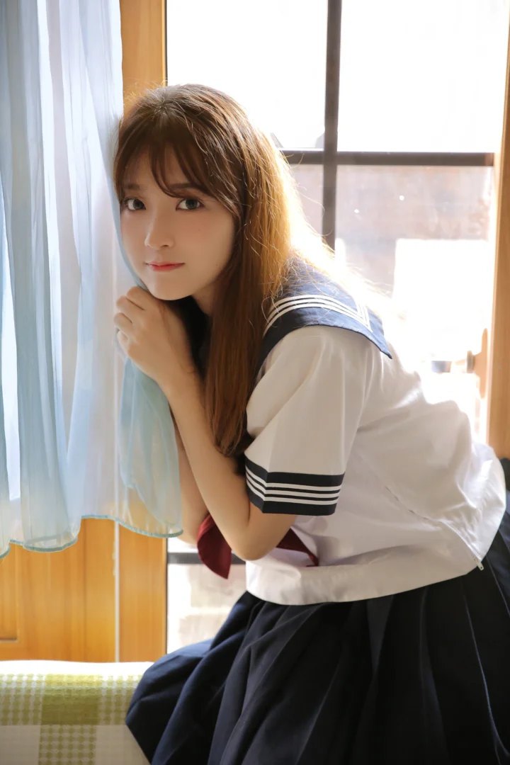 セーラー服風の衣装を着た成人女性が、窓辺で日差しを受けながら座っています。