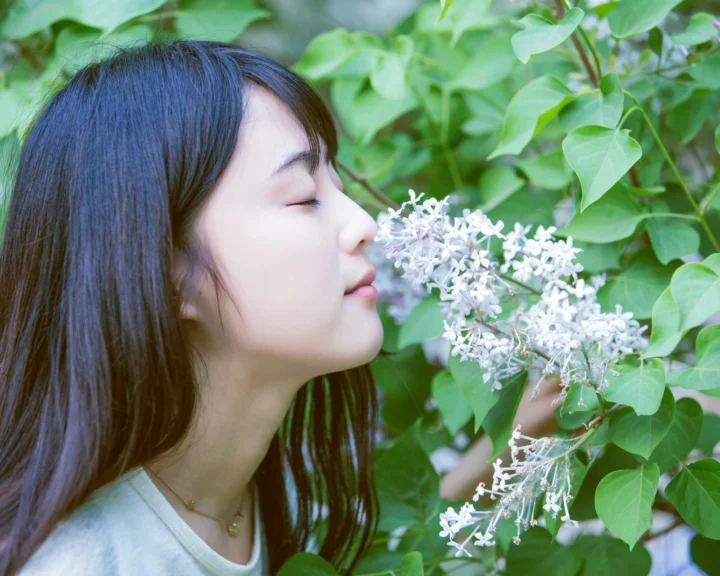 一名成年女性闭眼靠近白色花簇，周围是茂密的绿色叶片。