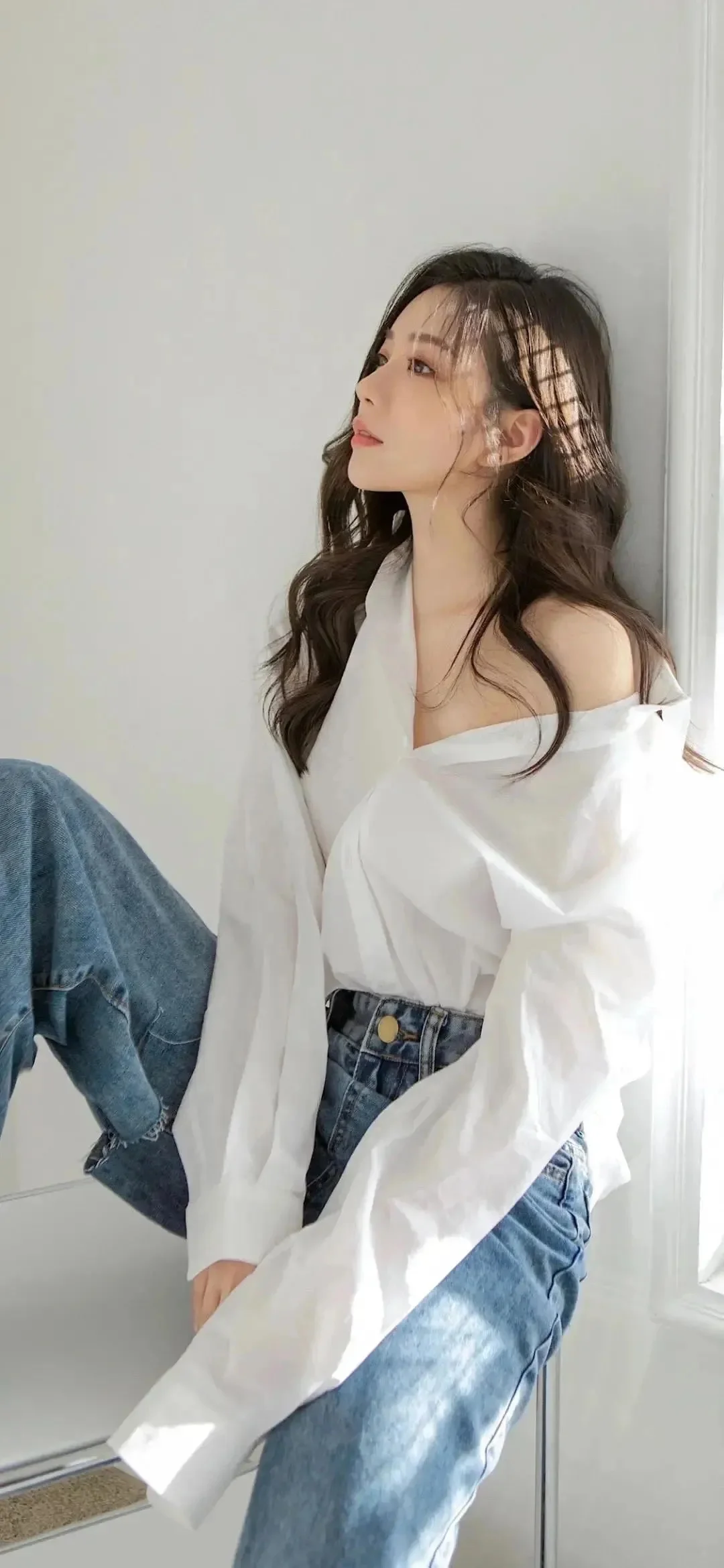 一名长发女子穿着白色衬衫和蓝色牛仔裤坐在室内窗边。