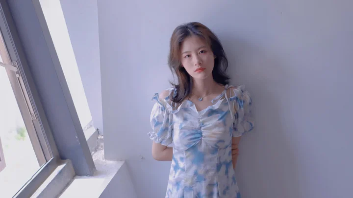 一名女子站在窗邊，穿著藍色連衣裙。