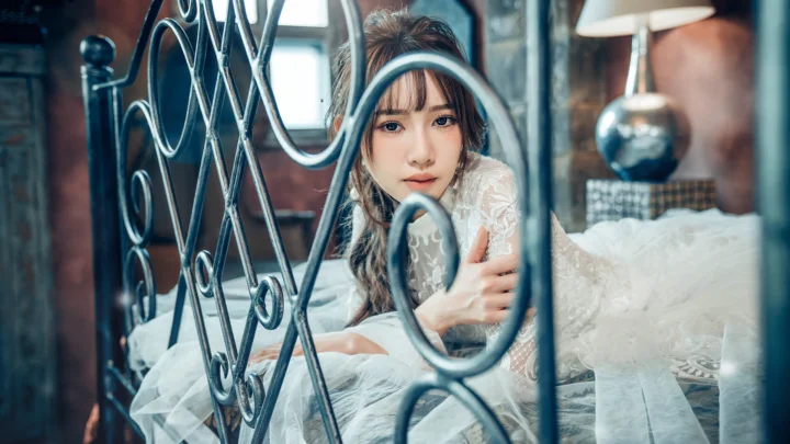 一名成年女子穿着白色蕾丝礼服，隔着装饰性金属床架望向镜头。