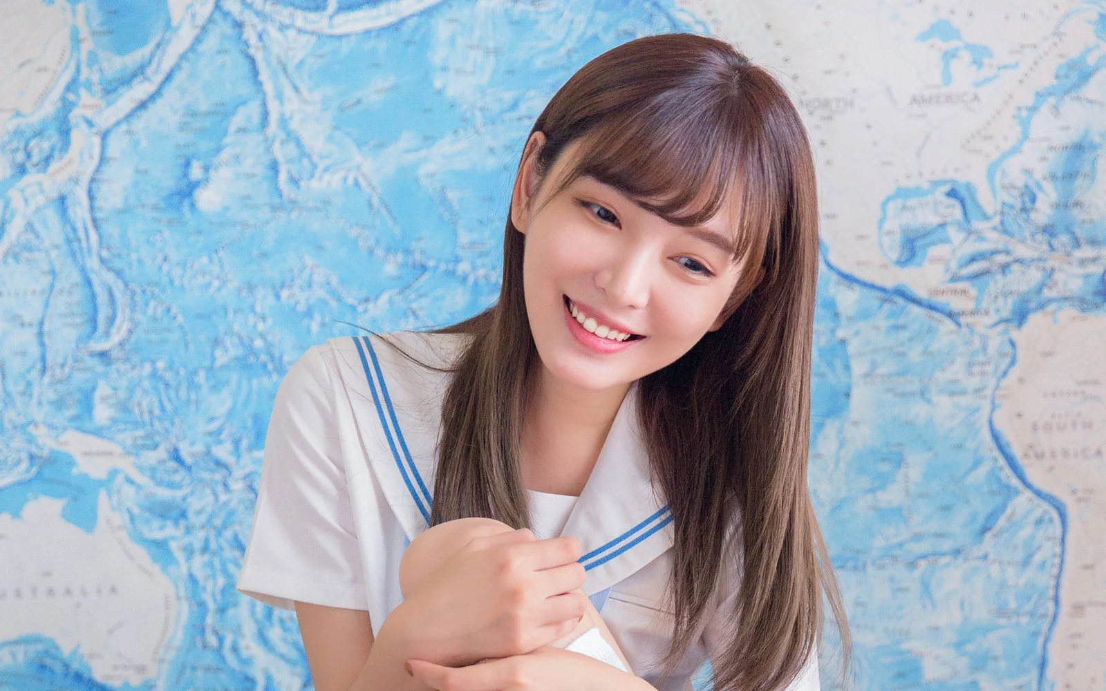 파란 지도 배경 앞에서 흰색 세일러 칼라 상의를 입은 긴 머리 여성이 미소 짓고 있다.