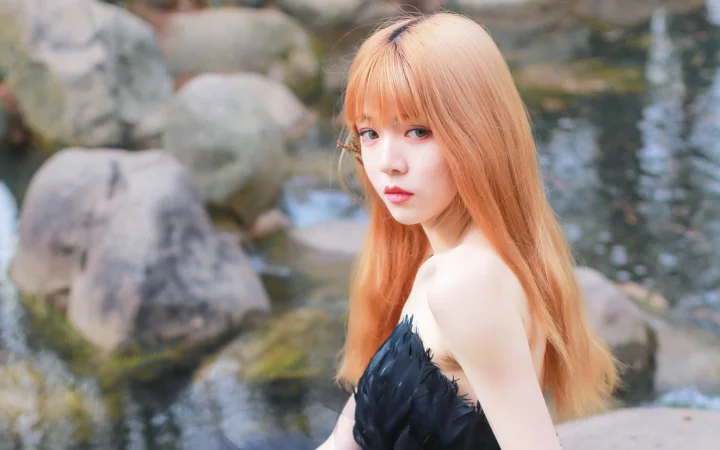 검은 드레스를 입은 빨간 머리 여성이 바위가 있는 개울가에서 카메라를 바라보고 있습니다.