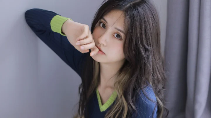 긴 머리의 여성이 실내 커튼 근처에서 카메라를 바라보며 인물 사진 포즈를 취하고 있다.