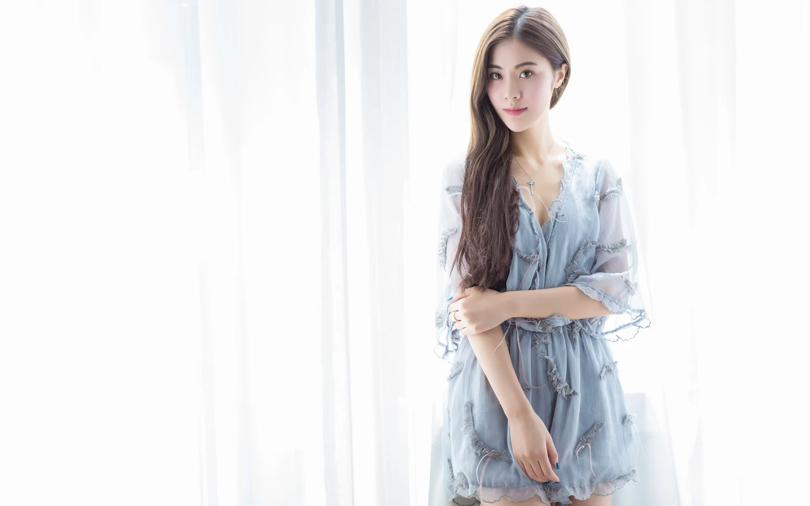 一名成年女子穿著淺藍色連身裙站在明亮窗簾旁。