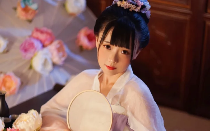 一名身穿浅色衣服、佩戴花饰的女子手持圆镜坐在室内，身后可见花束和木质背景。