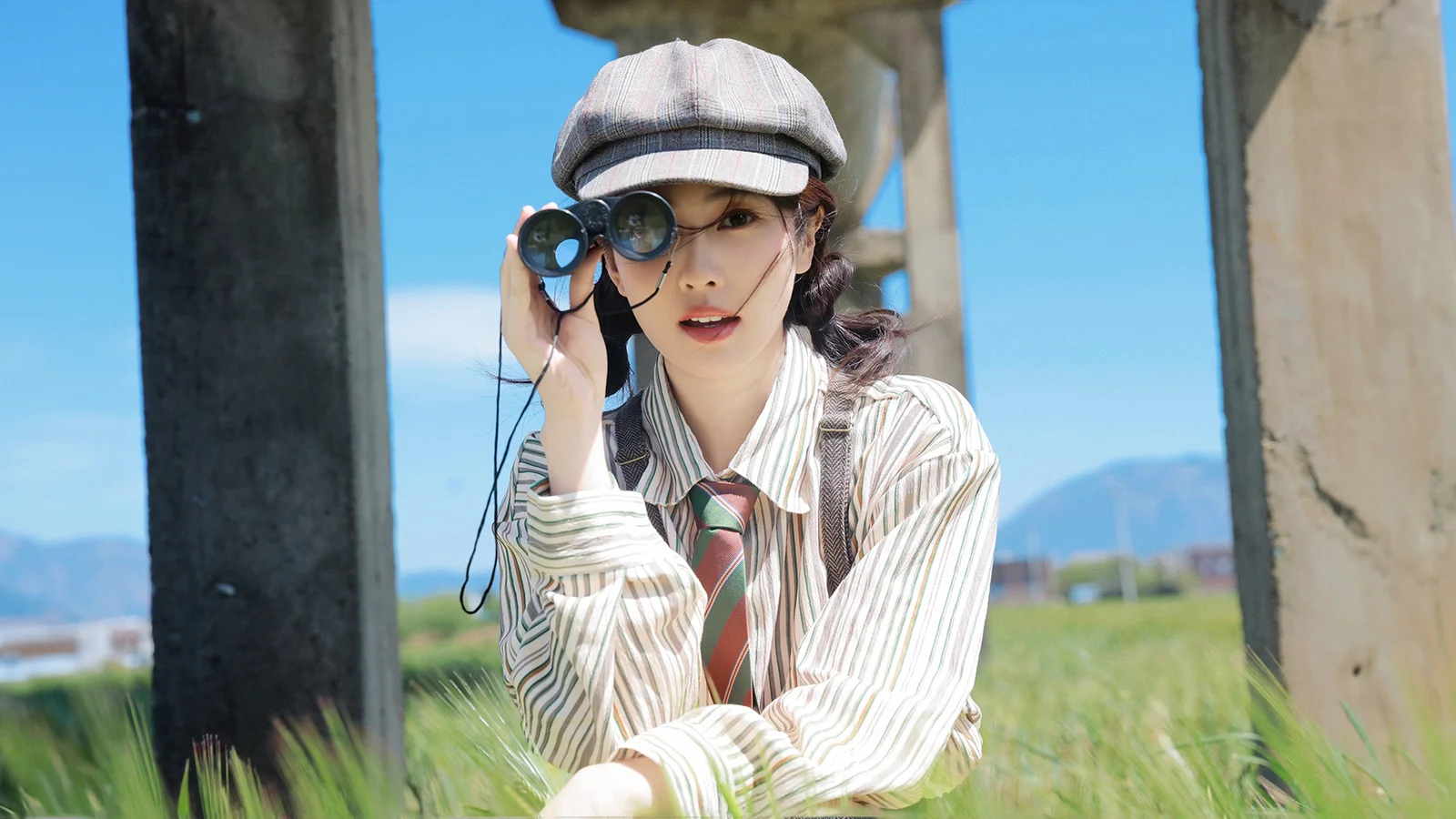 一名戴帽子的成年女性在草地上手持望遠鏡，身後是藍天、山景和混凝土柱子。