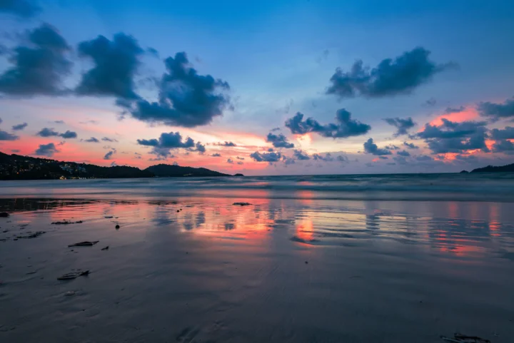 夕阳映照着海滩与平静海面，云层和晚霞倒映在潮湿的沙滩上。