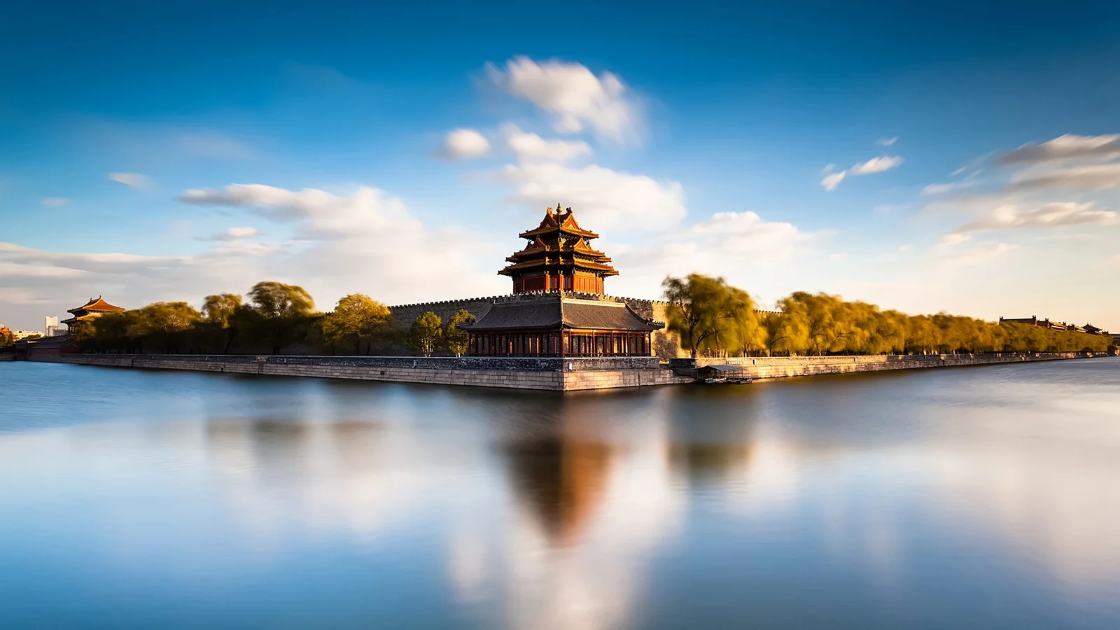 Landscape desktop wallpaper of a traditional Chinese palace corner tower by a still moat, with walls, trees, and a blue sky reflected in the water.