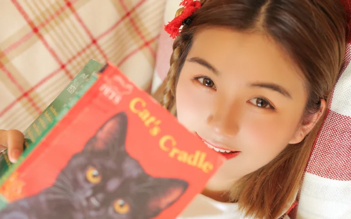 一名女子近距离看向镜头，手中举着一本印有黑猫图案的书。