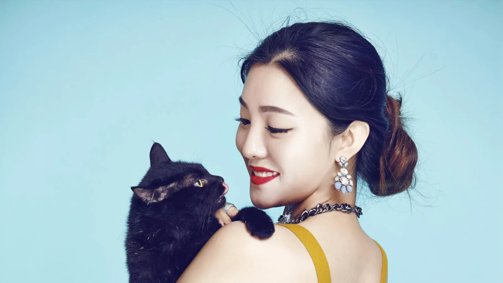 한 여성이 연한 파란색 배경 앞에서 검은 고양이를 안고 미소 지으며 바라보고 있다.