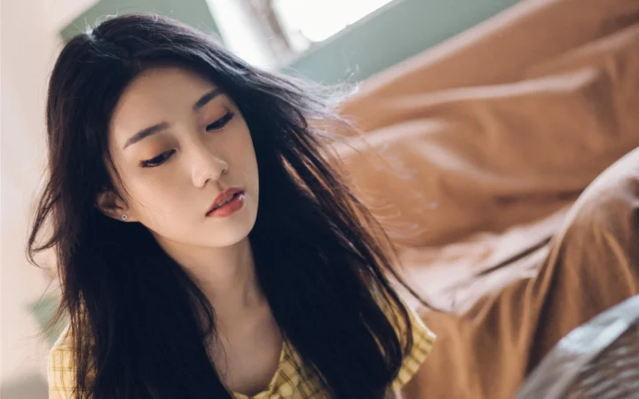一名黑发女性坐在室内，背景是柔和光线下的床铺或沙发。