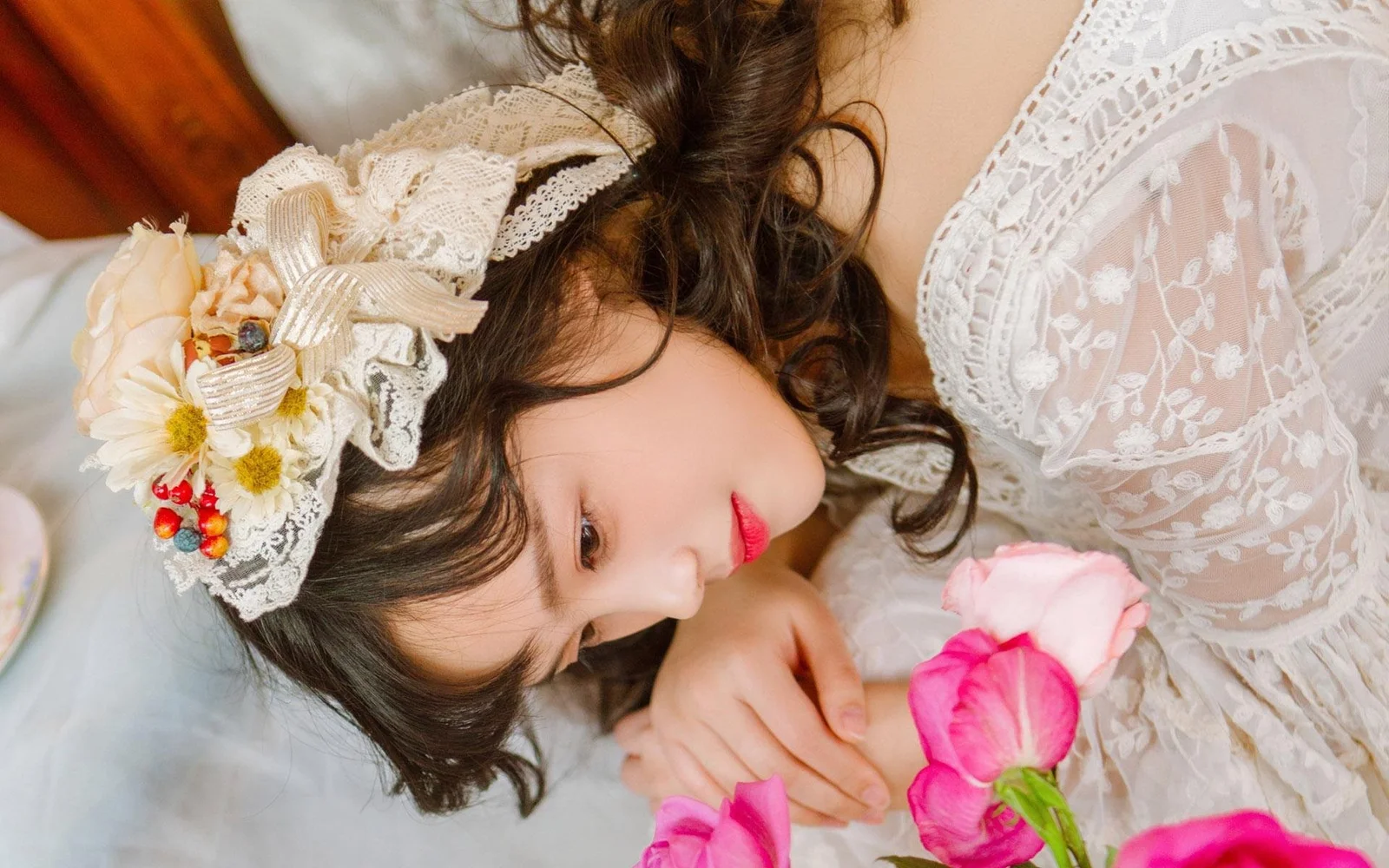 一位戴着花饰、身穿白色蕾丝服装的女性侧卧在床上，手边摆放着粉色花朵。