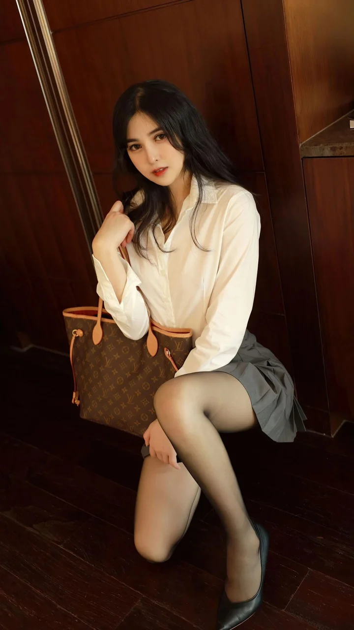 흰색 블라우스와 회색 치마를 입은 성인 여성이 나무 바닥의 실내에서 갈색 핸드백을 들고 앉아 있습니다.