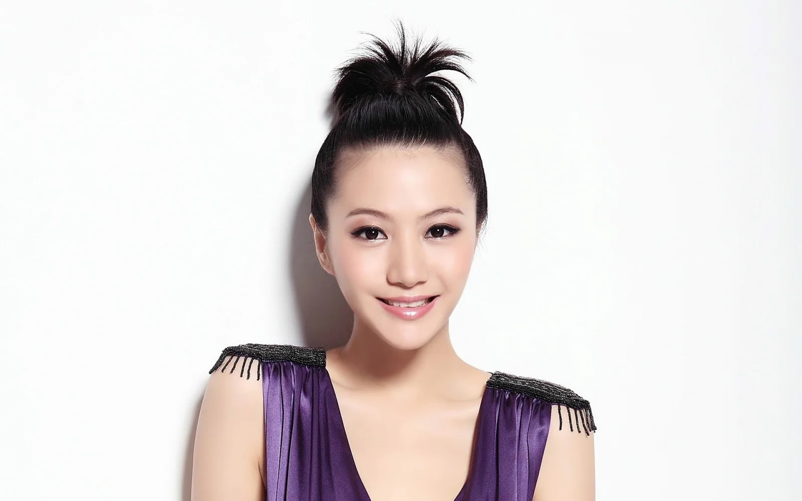 一名身穿紫色服装、盘发并面带微笑的女性站在白色背景前。