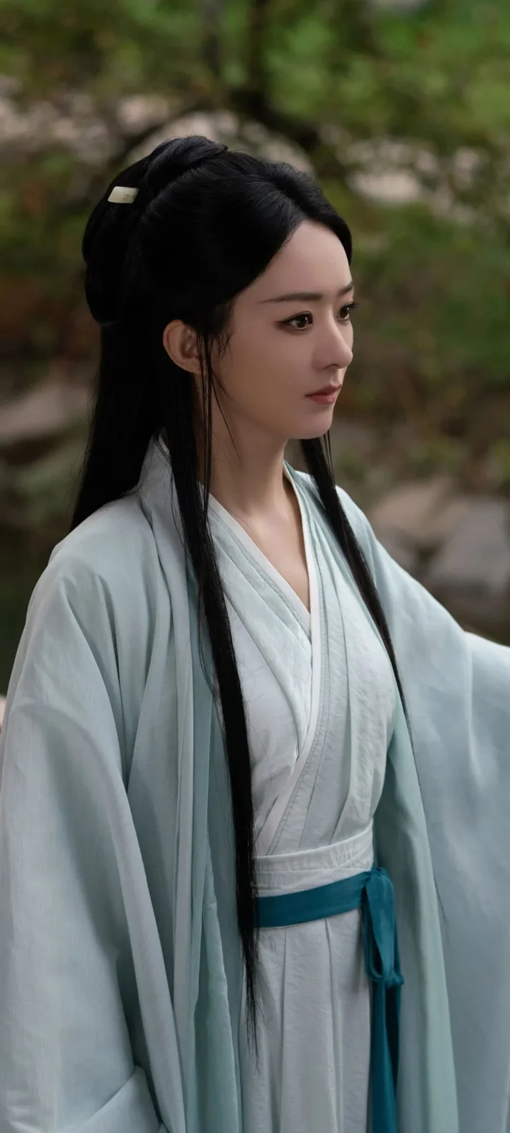 긴 검은 머리의 여성이 연한 파란색 전통 의상을 입고 야외의 푸른 식물을 배경으로 서 있습니다.
