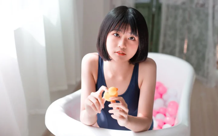 一名短发女子坐在白色浴缸中，手里拿着一个橙黄色物体，背景可见粉色物品。