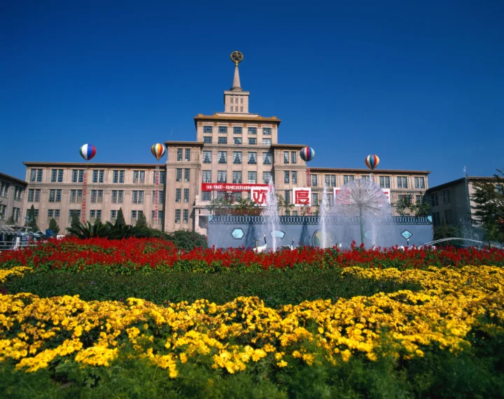 一座对称的城市建筑矗立在盛开的红黄花园后方，前方可见喷泉和彩色气球。