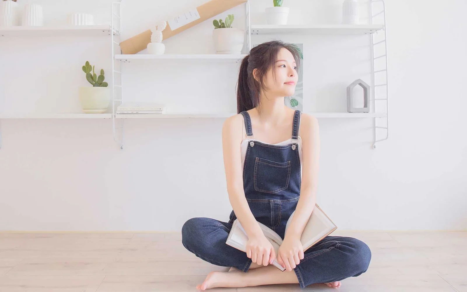 一名成年女性穿著牛仔吊帶褲坐在明亮的室內地板上，手中拿著一本書。
