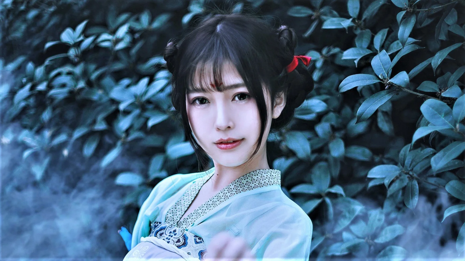 연한 파란색 전통풍 의상을 입은 여성이 짙은 녹색 잎을 배경으로 서 있습니다.