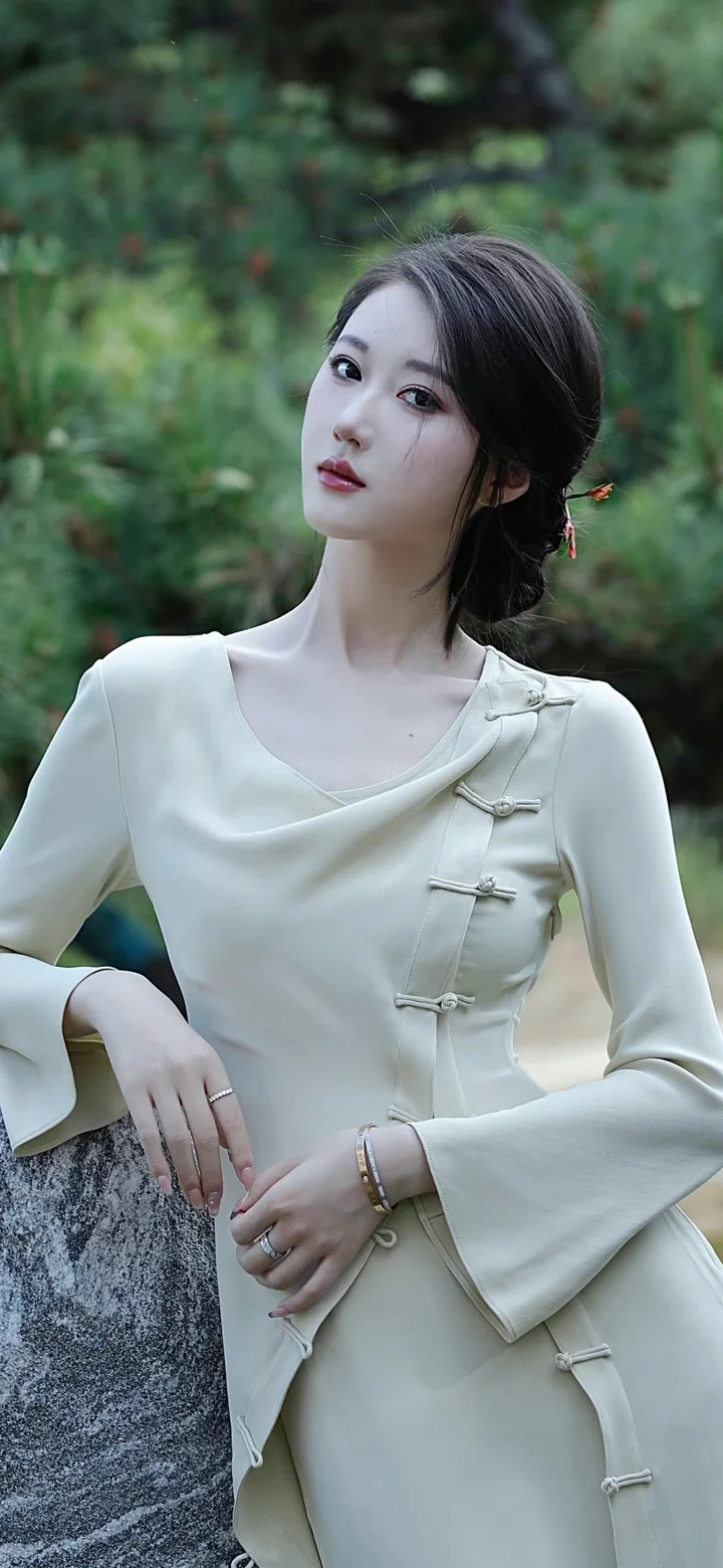 一名成年女性身穿浅色带盘扣的服装，在绿叶背景前倚靠石栏凝视镜头。