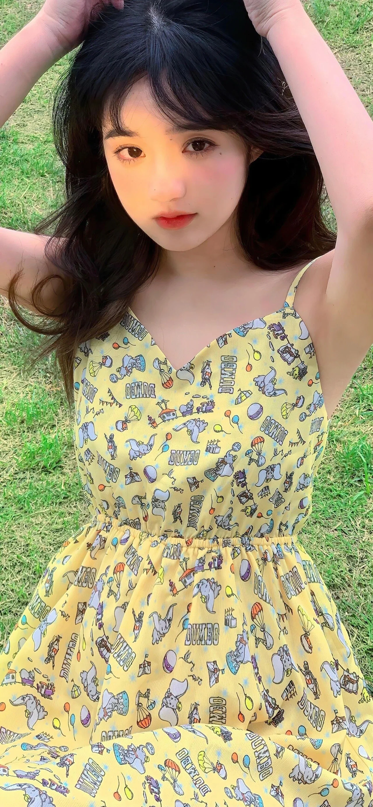 검은 머리의 여성이 무늬가 있는 노란색 원피스를 입고 잔디 위에 앉아 양팔을 머리 위로 올리고 있습니다.