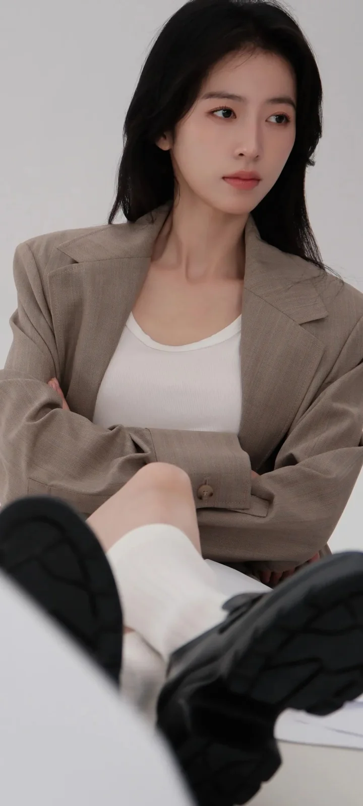 一名成年女性穿着棕色西装外套和白色上衣坐在浅灰色背景前，双腿穿着白袜和黑色靴子。